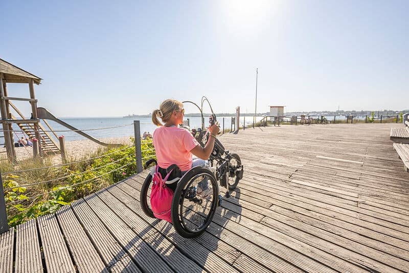 Person im Rollstuhl faehrt auf einem breiten Holzsteg am Strand und nutzt ein Handbike; links ist Meer und Sandstrand, das Licht ist sonnig.