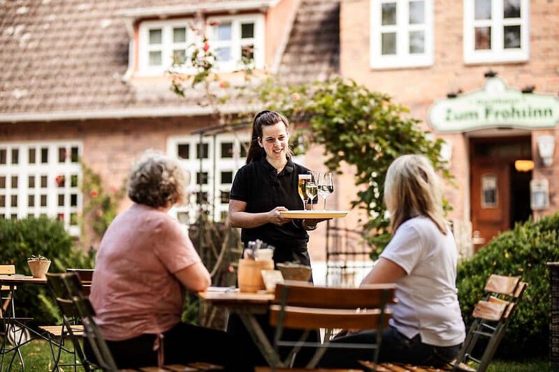 Eine Kellnerin trägt im Biergarten ein Tablett mit drei Weingläsern zu zwei sitzenden Gästen; im Hintergrund ein Backsteingebäude mit dem Schild „Zum Frohsinn“.