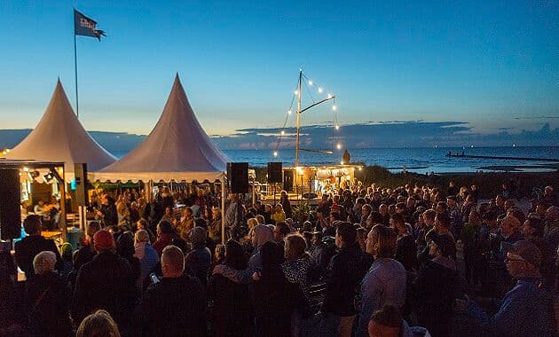 Große Menschenmenge bei einer Abendveranstaltung am Meer, vor zwei weißen Zeltdächern mit Bühne und Lichterketten.