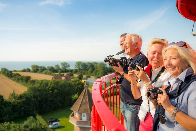 Vier ältere Personen stehen auf einer Aussichtsplattform mit rotem Geländer, halten Kameras und lächeln in die Ferne; im Hintergrund Felder, Bäume und das Meer am Horizont.