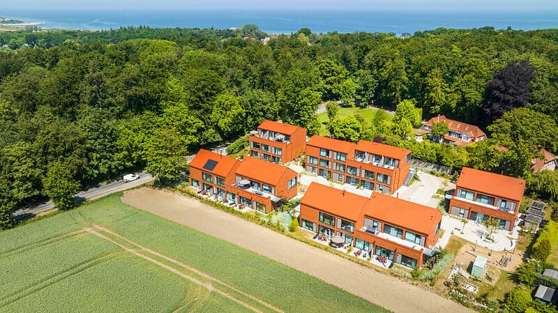 Luftaufnahme einer Wohnanlage mit mehreren modernen Häusern mit roten Dächern am Waldrand; dahinter ist das Meer zu sehen.