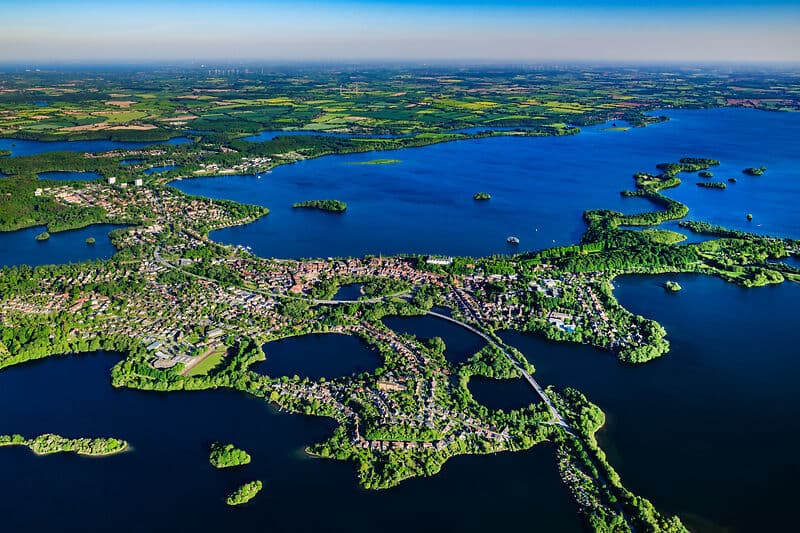 Luftaufnahme einer Seenlandschaft: blaue Seen mit vielen Buchten und kleinen Inseln, dazwischen eine Stadt mit Brücken und grüne Felder bis zum Horizont.