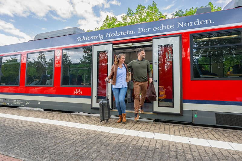 Ein Paar steigt am Bahnsteig aus einem roten Zug aus; die Frau zieht einen Rollkoffer. Auf dem Zug steht „Schleswig-Holstein. Der echte Norden.“