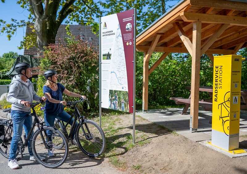 Zwei Radfahrende mit Helmen halten neben ihren Fahrrädern und lesen eine Infotafel „Willkommen am Ochsenweg“; daneben stehen ein Holzunterstand und eine gelbe „Radservice Station“.