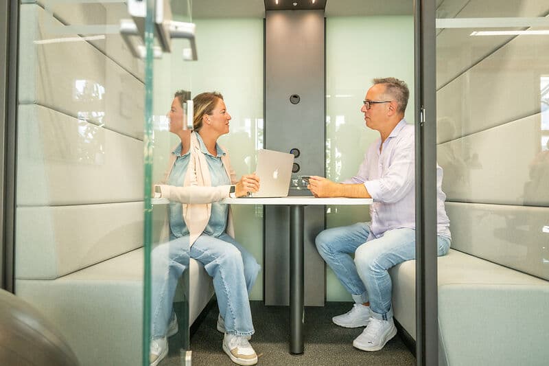 Zwei Personen sitzen sich in einer verglasten Büro-Box an einem kleinen Tisch gegenüber und sprechen; auf dem Tisch steht ein geöffneter Laptop.