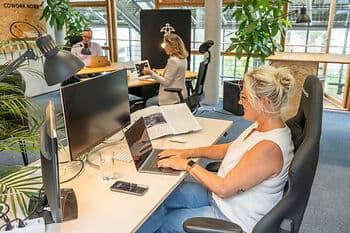 Blick von oben: Eine Frau sitzt in einem Strandkorb im grünen Garten und arbeitet am Laptop; neben ihr steht ein kleiner runder Tisch mit Smartphone.
