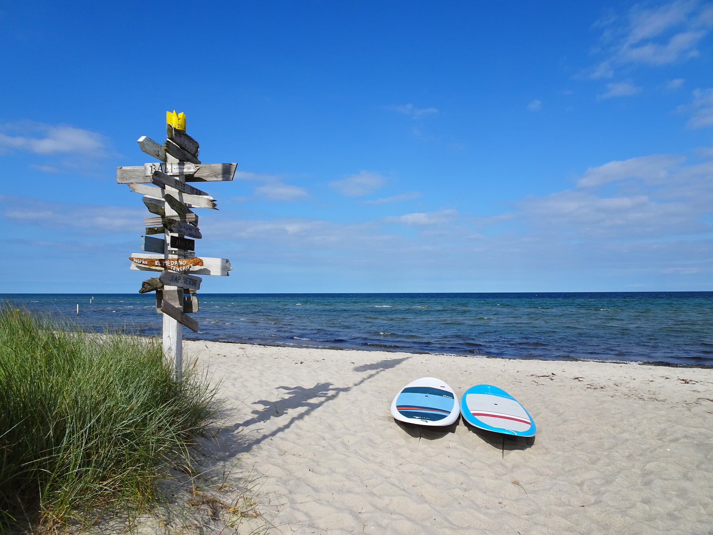 Sandstrand mit blauem Himmel und ruhigem Meer; links steht ein schiefer Wegweiser aus vielen Holzschildern, rechts liegen zwei Surfboards im Sand.