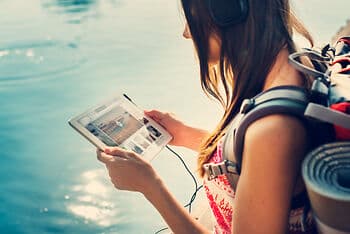 Frau mit Rucksack und großen Kopfhörern steht am Wasser und hält ein Tablet in beiden Händen.